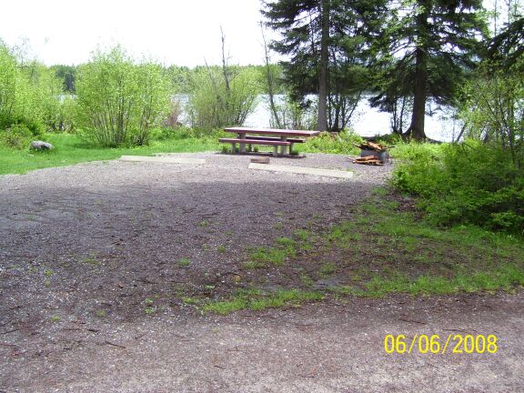 picture showing Two campsites with a large, shared, parking pad allowing two mobile homes to park side-by-side.  The lake can be seen through a strip of trees and shrubs behind the campsites. 