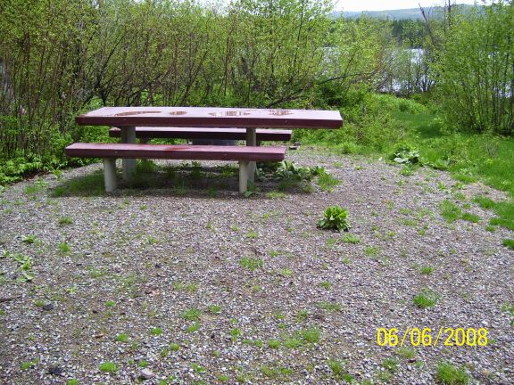 picture showing Every campsite has a picnic table similar to this one.  The picnic table is large, very sturdy, and has an extended tabletop providing a wheelchair seating space.  The table is permanently positioned on a gravel pad that extends from the campsite parking pad.  Notice that while the picnic table pad is level, it it does slope away away from the wheelchair seating position at the picnic table.  The slope (3.5 degrees) is most pronounced at this particular table.