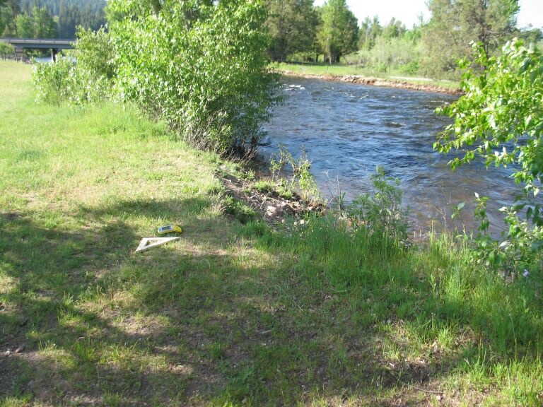 picture showing One of the better river access points showing a flat uneven grassy field (the access route) and a 30-degree slope down the 3-foot high soft, unstable river bank composed of dirt and boulders.