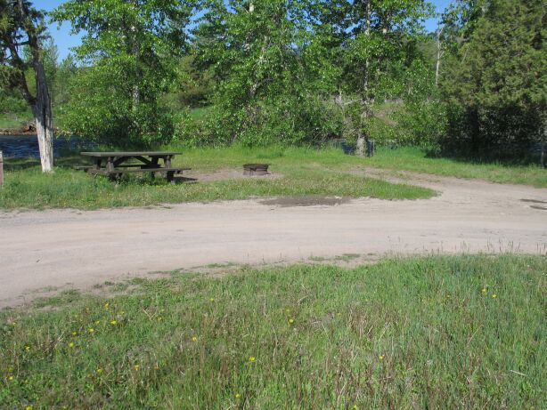 picture showing One of the more level campsites showing a picnic table without a wheelchair seating space, a BBQ pit with a cooking surface lower than 15 inches, and unevenly packed dirt and grass access routes.