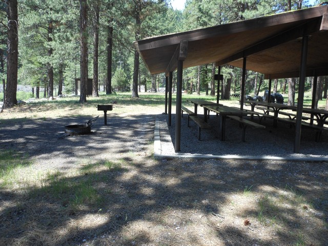 picture showing Picnic shelter with latrine in the far background.  There is a concrete curb around the perimeter of the shelter with loose gravel on the inside.  Best access to an accessible table would be on the far side of the shelter.