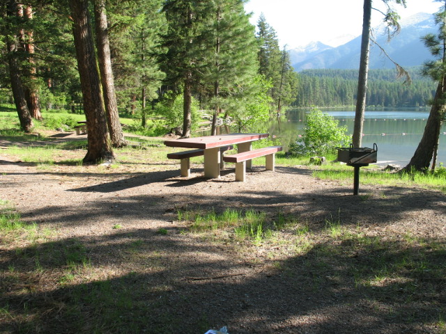 picture showing Day Use Area: One of three accessible picnic tables with a wheelchair seating space, BBQ stand, and firm and stable, although slightly uneven, gravel access route.  The dedicated swimming area and long sloping beach lawn can be seen in the background.