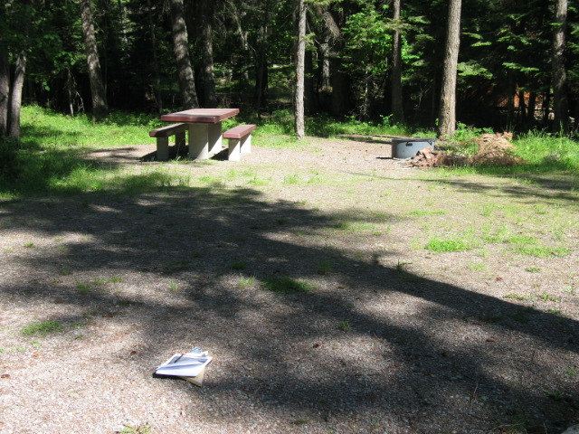 picture showing Most campsites are accessible.  This campsite shows a continuous, firm and stable, but slightly uneven in places, gravel surface extending from the parking pad around the picnic table and BBQ pit.