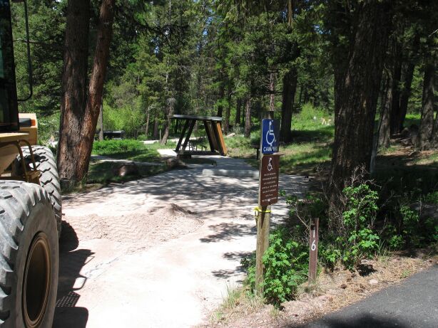 picture showing Accessible campsite (campsite number 15) showing paved parking pad and paved access route to a covered shelter with wheelchair-friendly picnic table and BBQ stand, and an electrical hookup.  The paved path can be seen continuing onto the accessible bathrooms and showers.