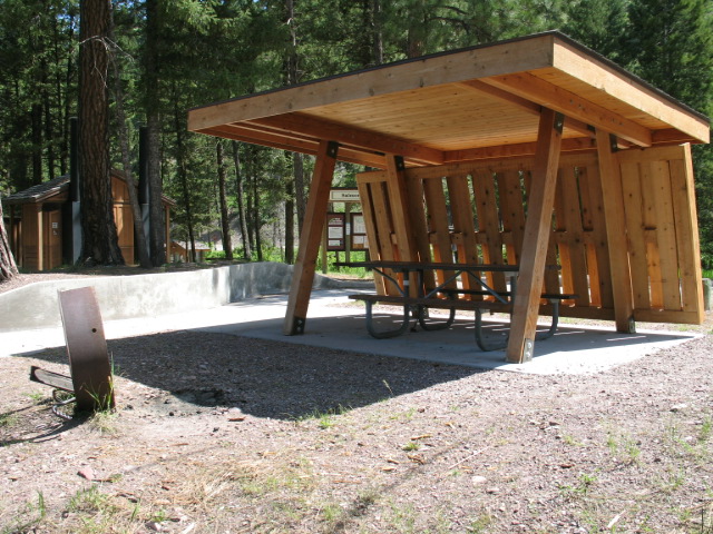 picture showing Sheltered accessible picnic area with an accessible picnic table and a paved access route from a paved, parking space.  Accessible bathrooms with pit toilets (also with a paved access route) can be seen in the distance.  The sheltered picnic area overlooks the lake.  This picnic area is located in southern day use area of the campground.