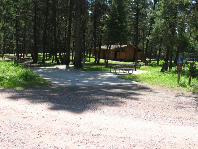 picture showing The accessible campsite in the campground.  A paved parking pad, wheelchair-accessible picnic table, and BBQ stand can be seen, as well as a paved path leading to a water spigot and an accessible bathroom and shower beyond the campsite.  This site has an electrical hookup.