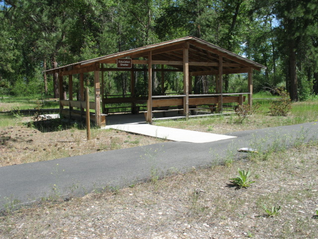 picture showing The Education/Picnic Shelter consisting of a large sheltered area lined with built-in benches with back support, and a large picnic table with a wheelchair seating space.  A concrete sidewalk connects the Education Center to the main paved path.