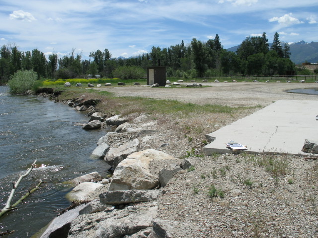 picture showing A 3.5-foot high rip rap river bank only a few feet from the gravel parking area.  An accessible bathroom can be seen in the distance, and the corner of a paved parking pad large enough for two vehicles (but not marked as accessible parking) can be seen to the right.  This is a very flat and open area.